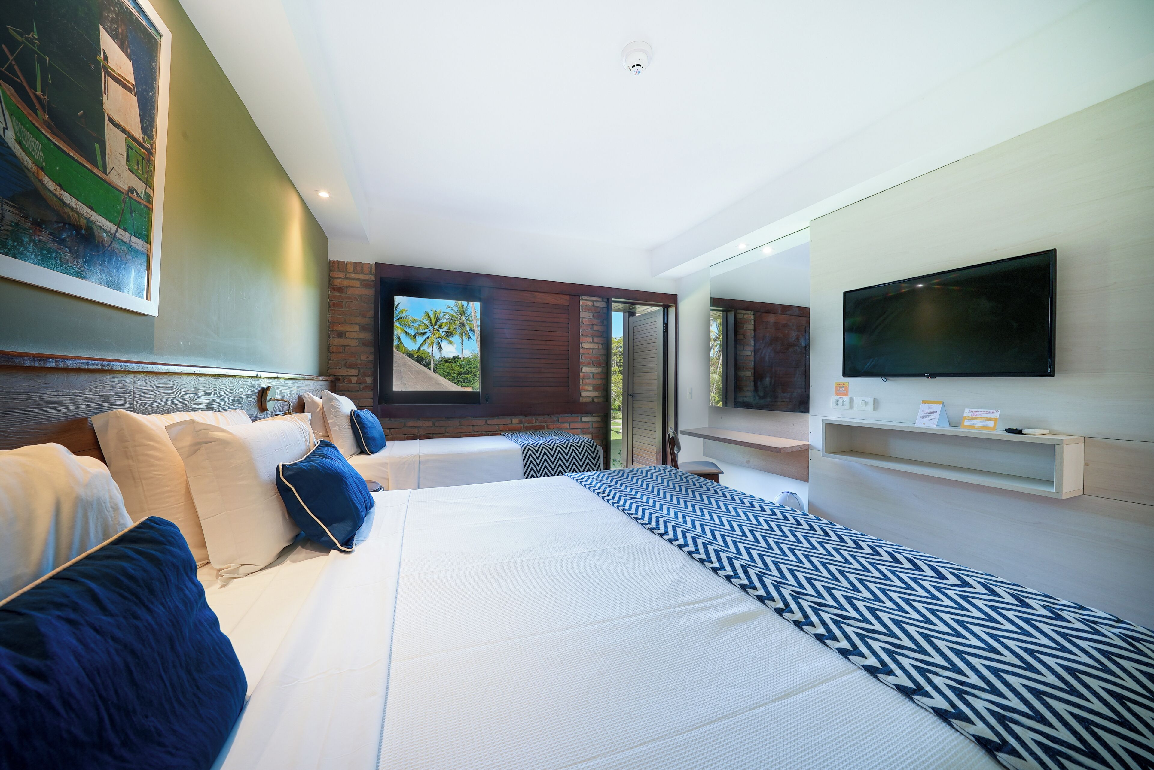 A modern hotel room with a large bed, a flat-screen TV, a painting, and a view of palm trees.