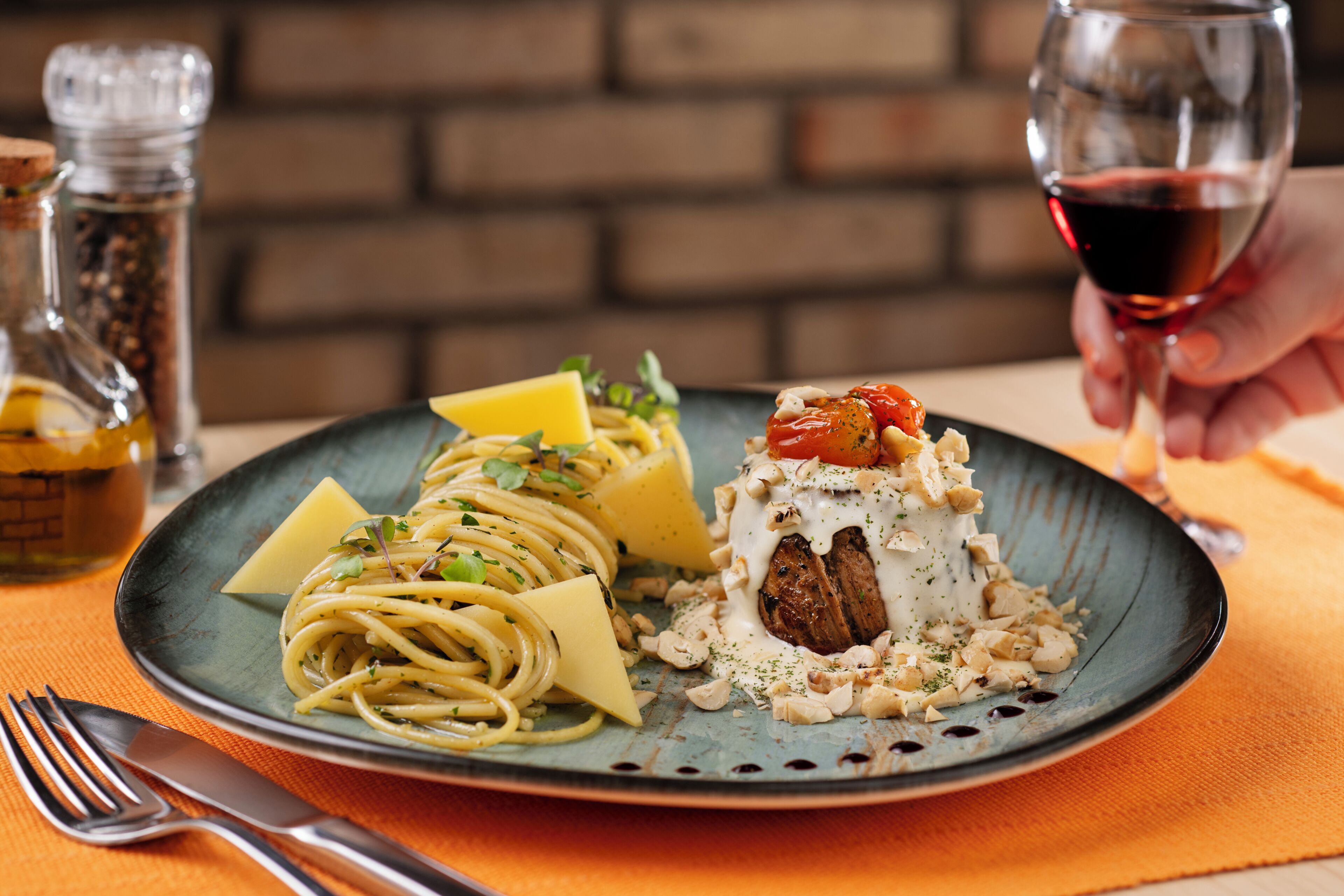 A plate of pasta with cheese and a meat dish, a glass of red wine, and a fork on an orange tablecloth.