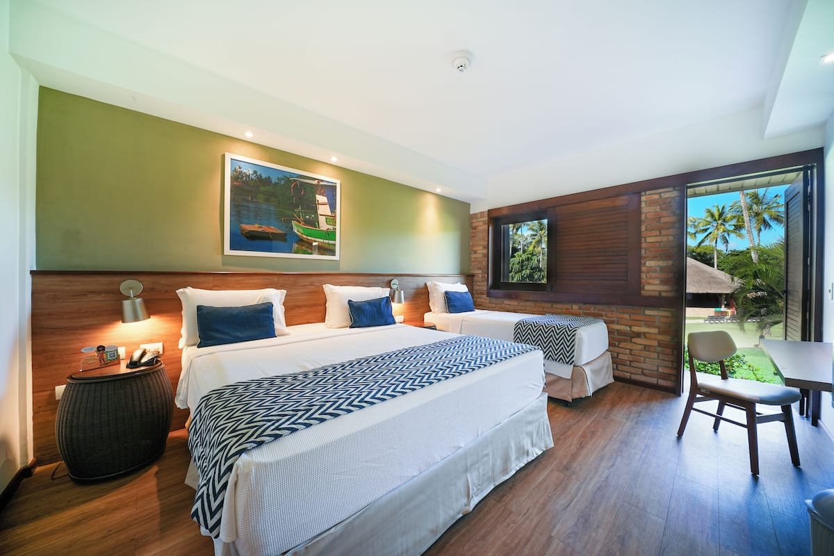 A hotel room with two beds, a desk, a chair, a TV, and a window with a view of palm trees.