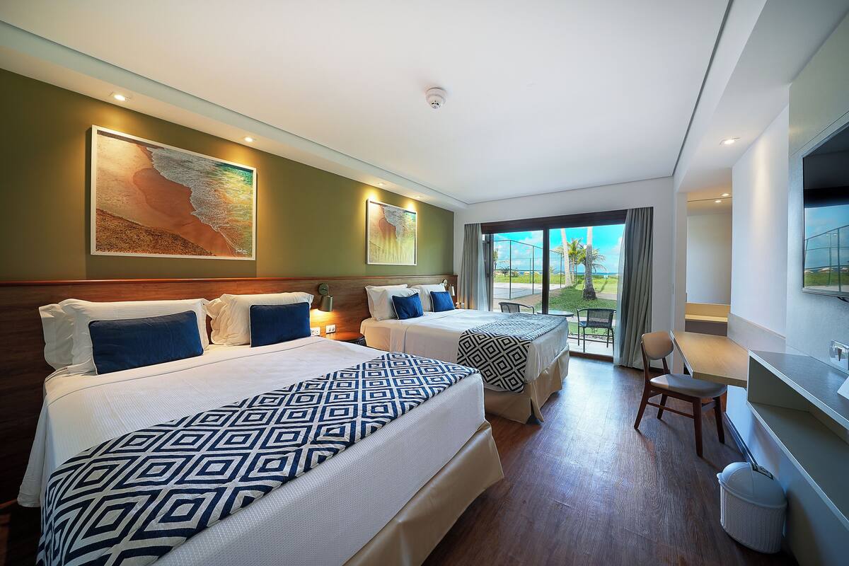 A hotel room with two beds, a desk, a chair, a TV, and a balcony with a view.
