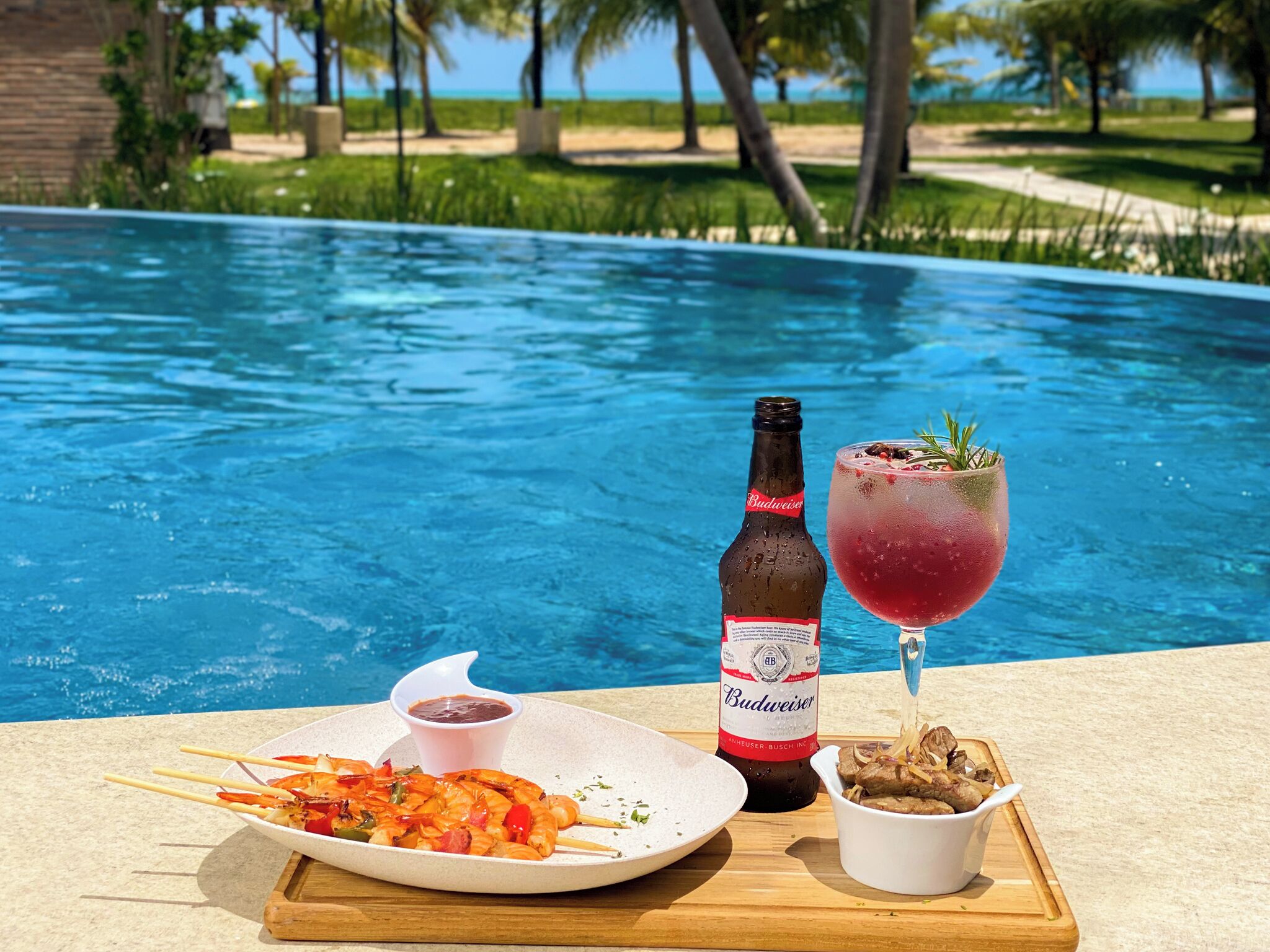 A poolside setting with a plate of shrimp skewers, a bottle of Budweiser beer, and a cocktail glass.