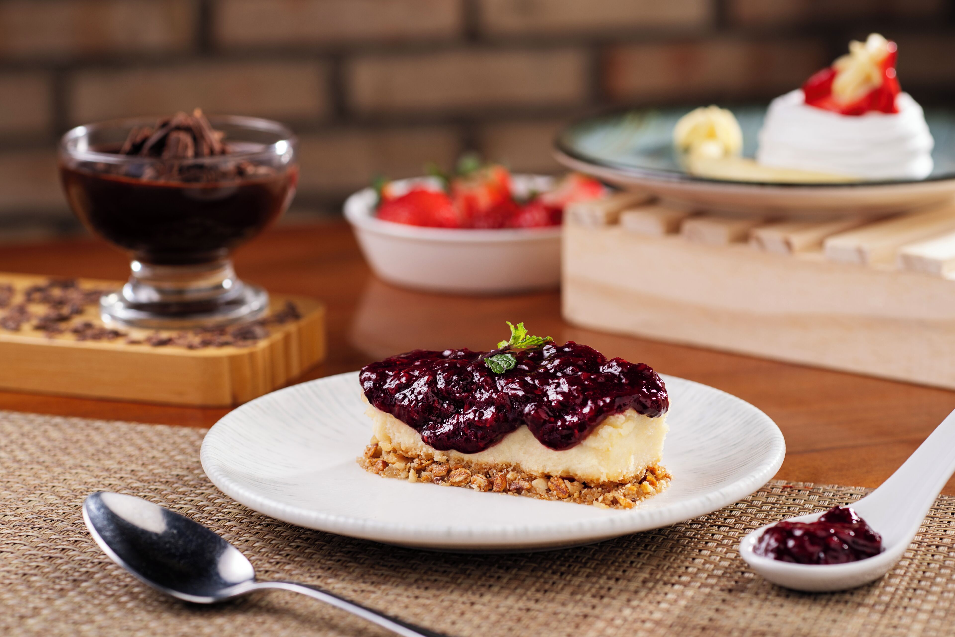 A dessert plate with a slice of cheesecake topped with berry compote, a bowl of chocolate sauce, and a bowl of strawberries.