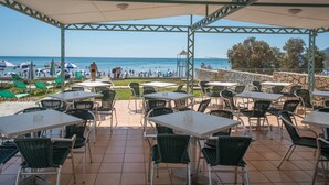 An outdoor patio area with tables and chairs, overlooking a beach with umbrellas and people. - Hotel Astir Palace (Zakynthos)