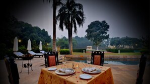 Un área de comedor al aire libre con una mesa dispuesta para dos, rodeada de sillas, palmeras y vistas a una piscina.