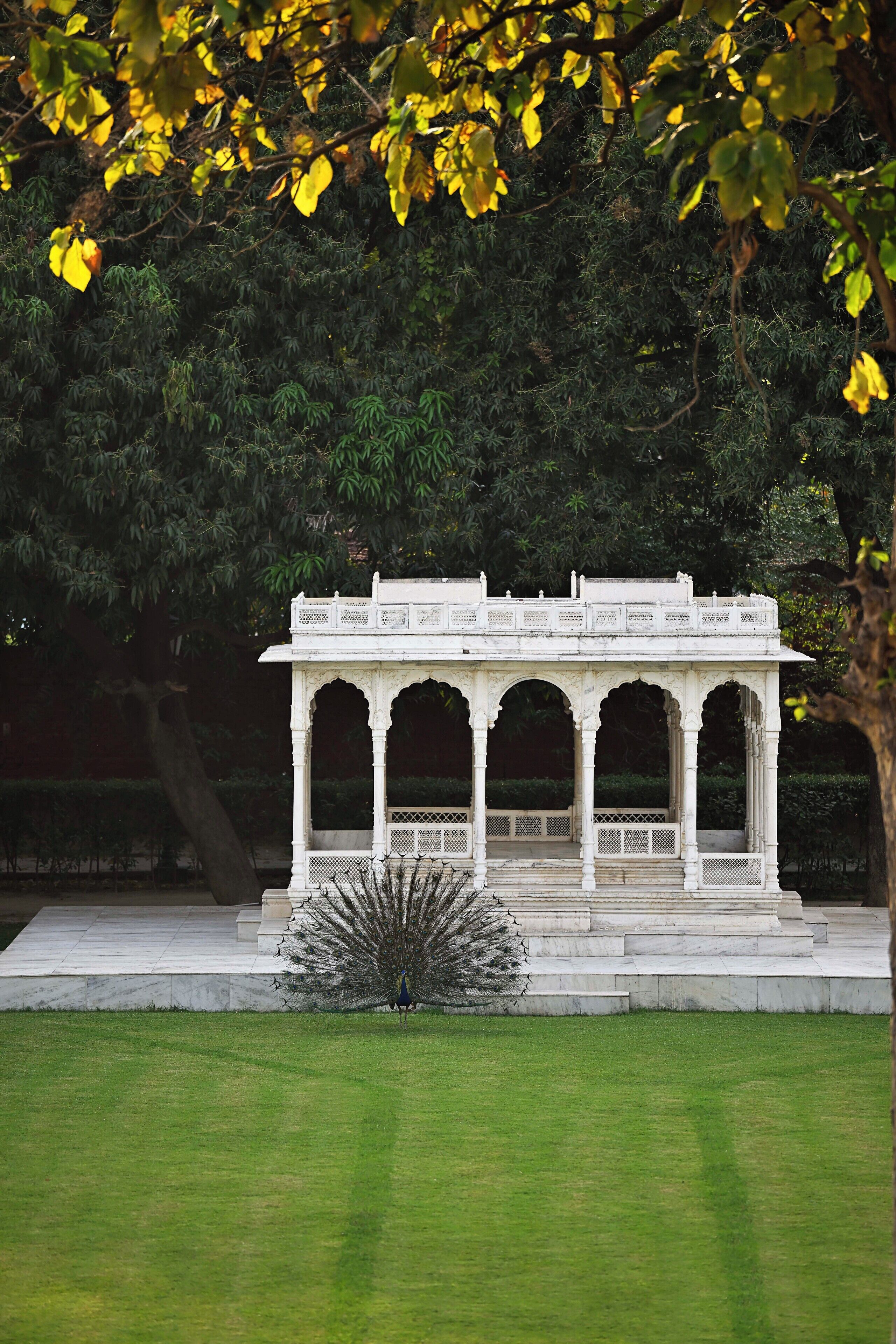 A white pavilion with arches and a central open space, surrounded by a well-maintained lawn and dense foliage.