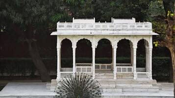 A white pavilion with arches and a central open space, surrounded by a well-maintained lawn and dense foliage.