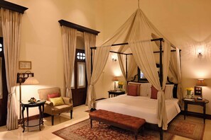 A four-poster canopy bed with a bench, a sitting area with a chair and table, and a nightstand with lamps. - Taj Nadesar Palace (Varanasi)