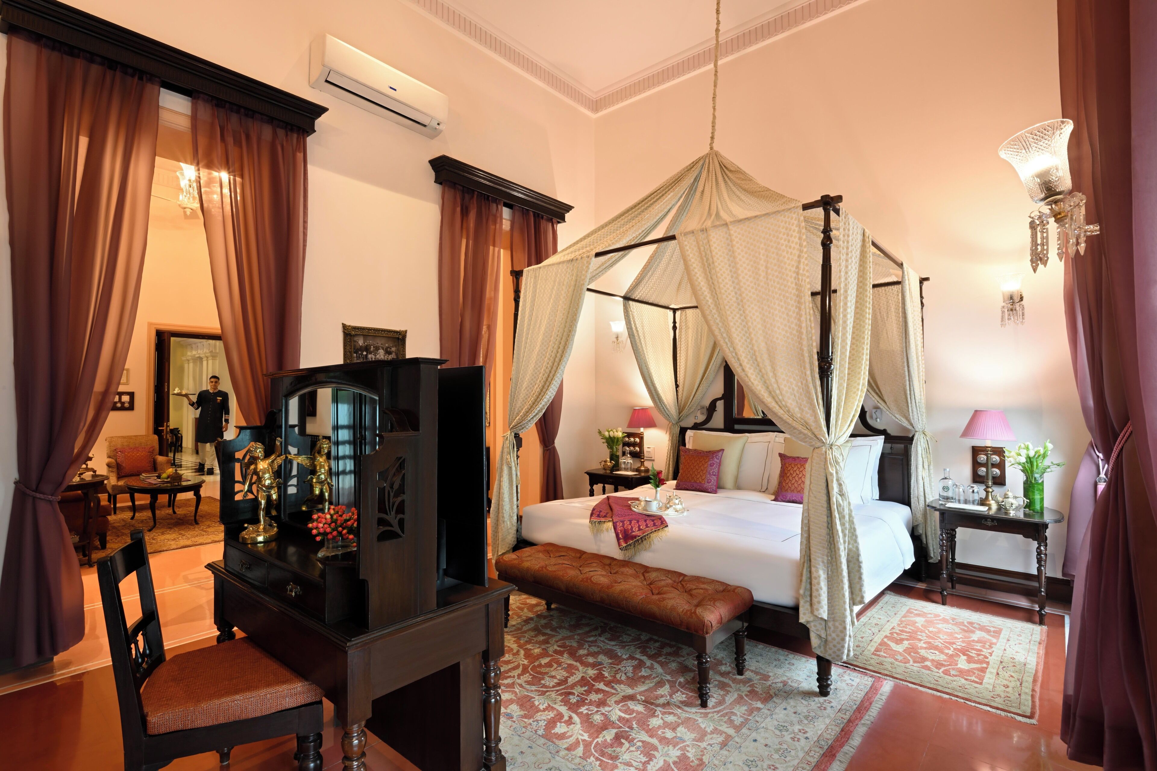 A four-poster bed with a canopy, a television on a wooden stand, a bench, and a small table with a lamp.