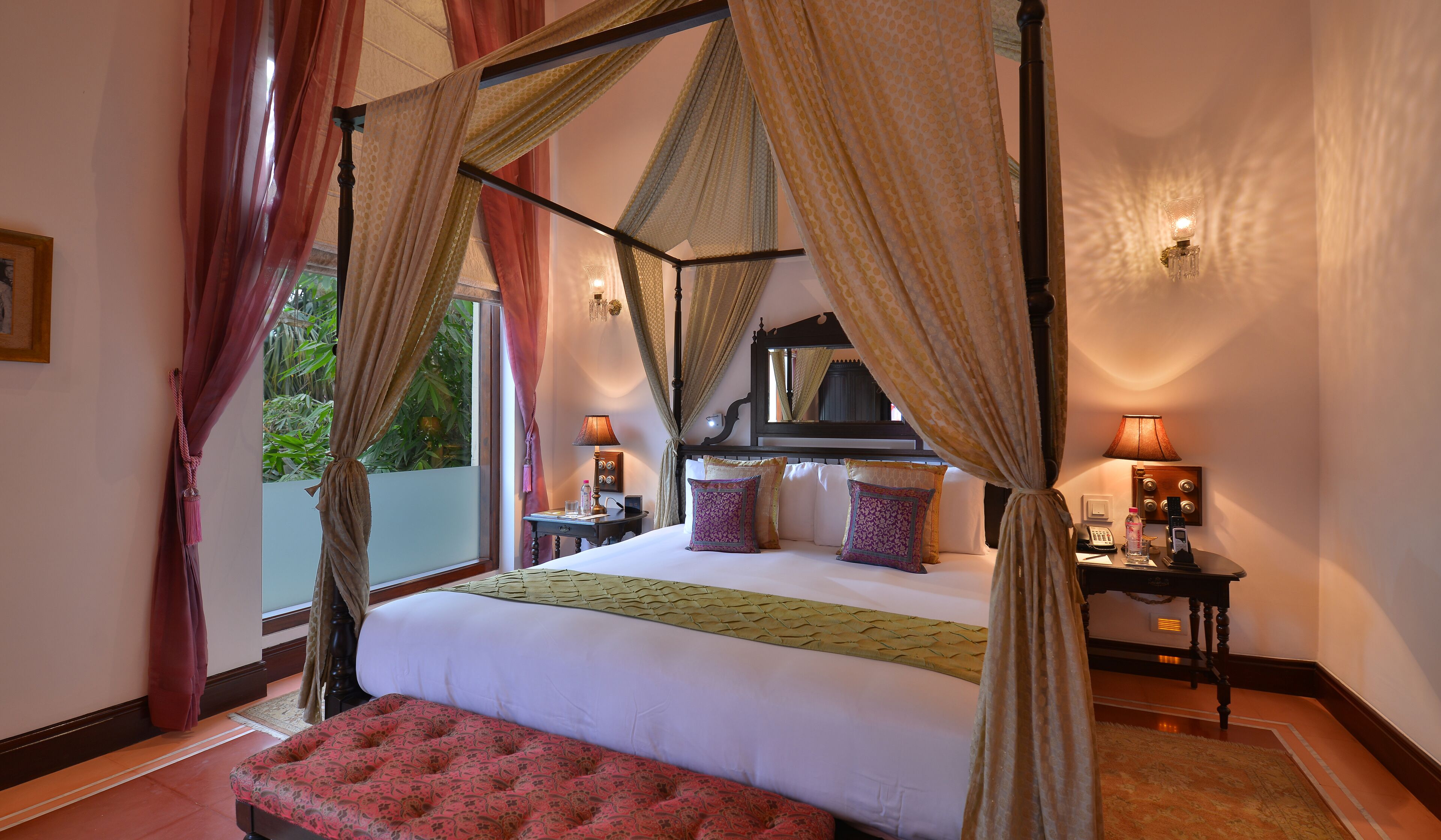 A four-poster canopy bed with a bench, bedside tables with lamps, and a view of greenery outside.