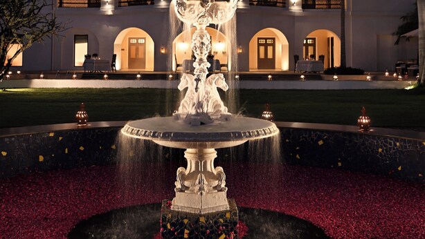 Una fuente con una estatua en el centro, rodeada por un parterre circular de flores con pétalos rojos. En el fondo, se observa un edificio grande con varios balcones y arcos.