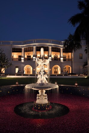 A fountain with a statue in the center, surrounded by a circular flower bed with red petals. In the background, there is a large building with multiple balconies and arched doorways. - Taj Nadesar Palace (Varanasi)