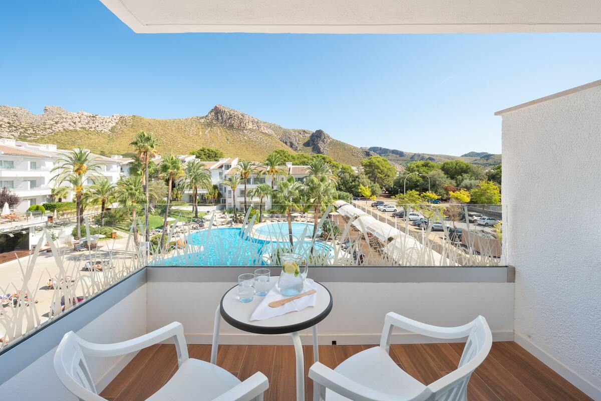 Un balcon avec deux chaises blanches, une petite table ronde et une vue sur une piscine et les bâtiments du complexe hôtelier.