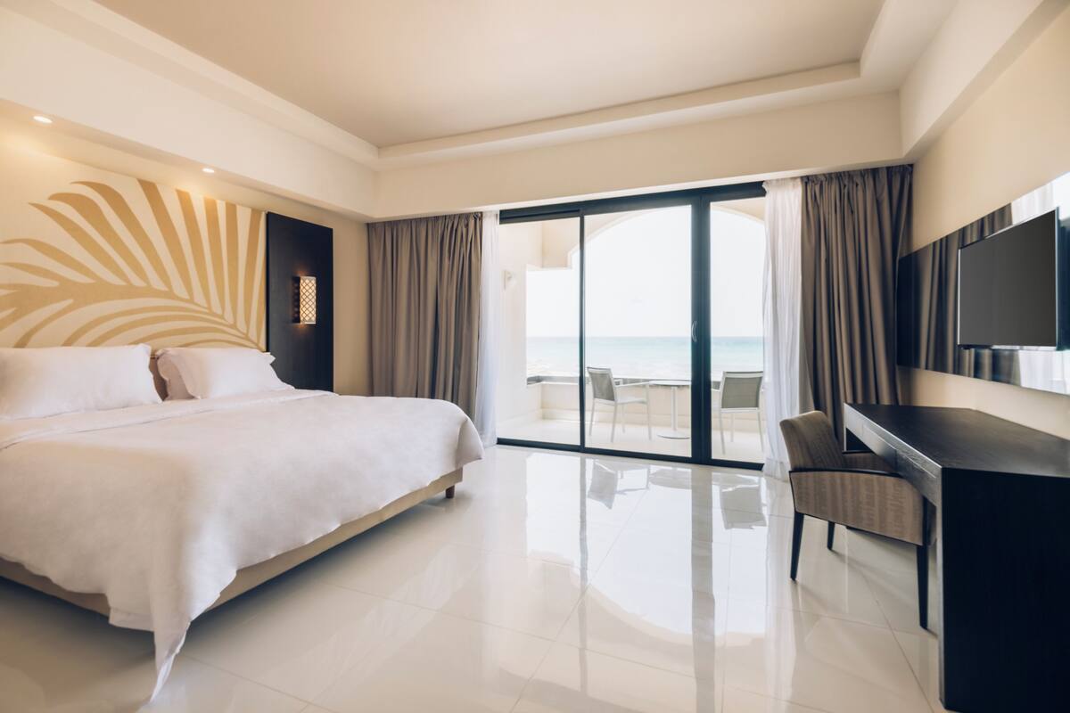 A modern hotel room with a large bed, a desk, a flat-screen TV, and a balcony with a view of the ocean.