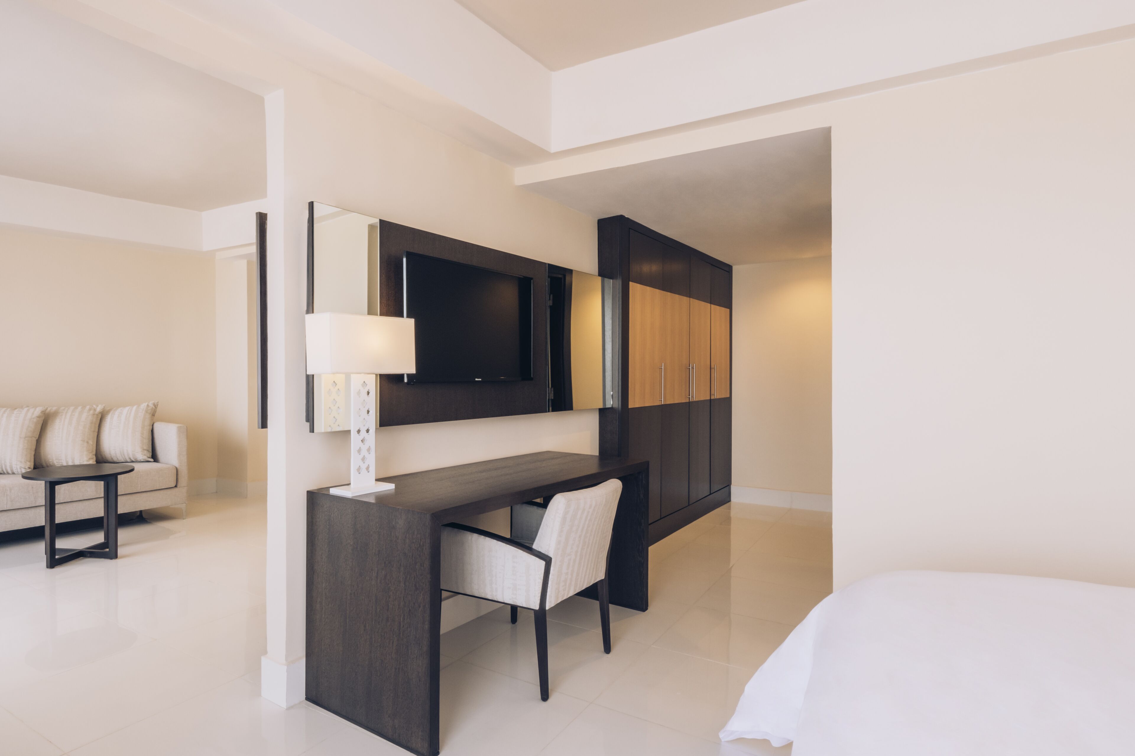 A modern hotel room with a dark wood desk, a chair, a flat-screen TV mounted on the wall, a sofa, and a bed.