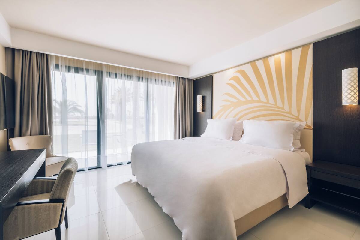 A hotel room with a large bed, a desk, and a chair. There is a large window with curtains, and a wall with a decorative panel.