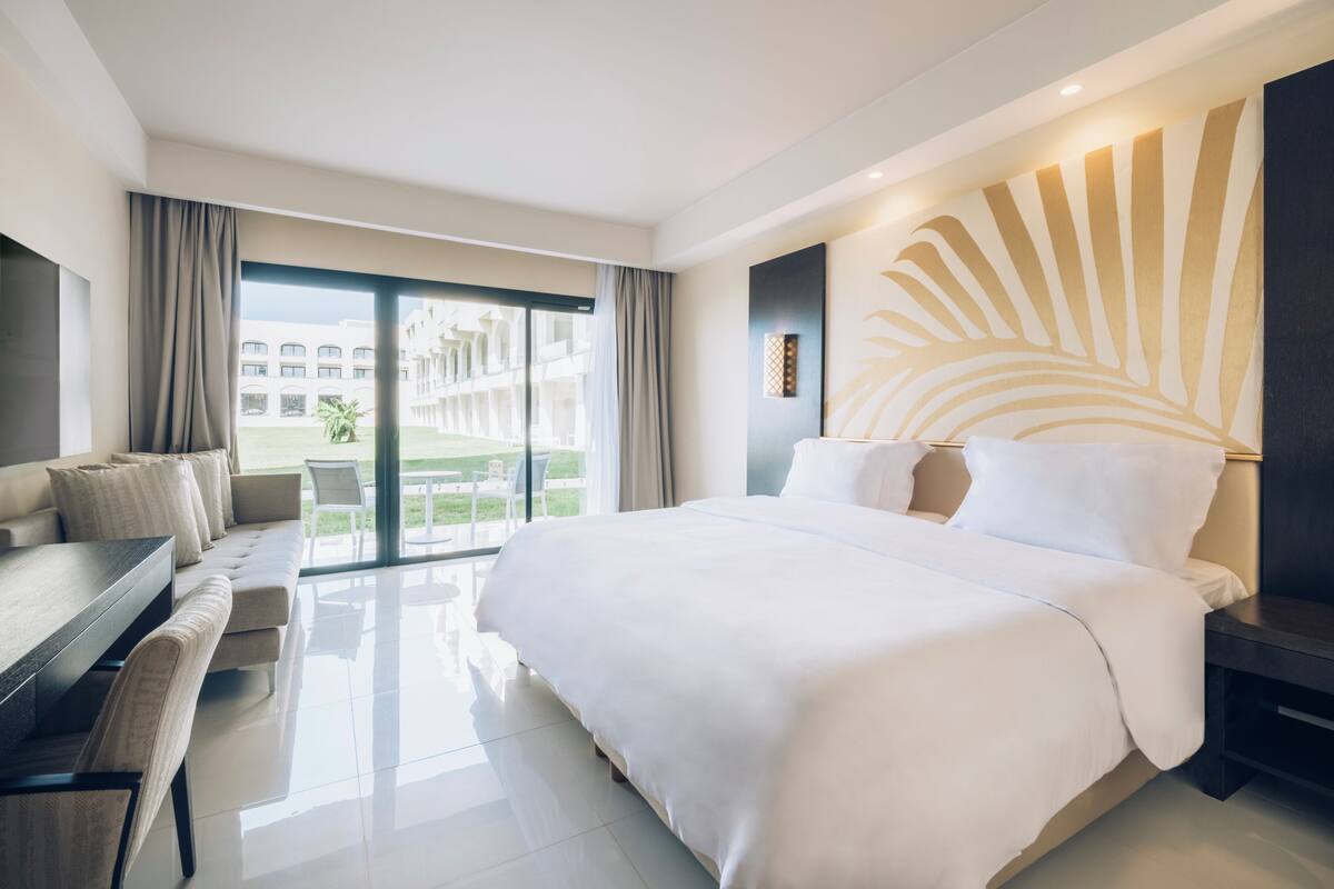 A modern hotel room with a large bed, a sofa, a desk, and a view of a building and greenery outside.