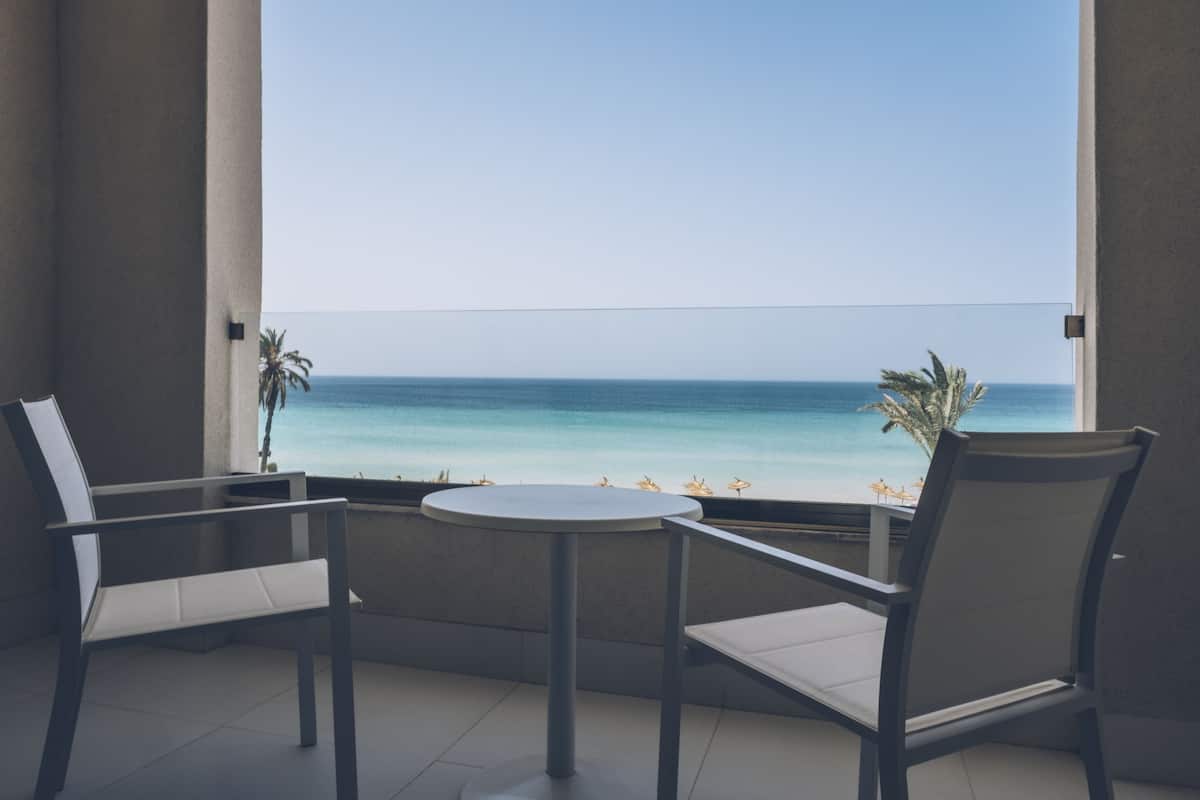 A balcony with two chairs and a small table, overlooking a beach with palm trees and the ocean.