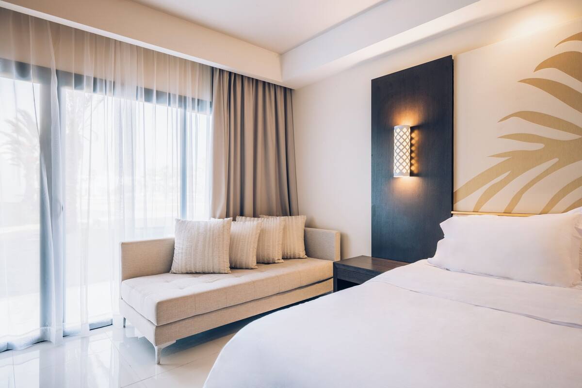 A modern hotel room with a large bed, a sofa, and a wall-mounted light fixture.