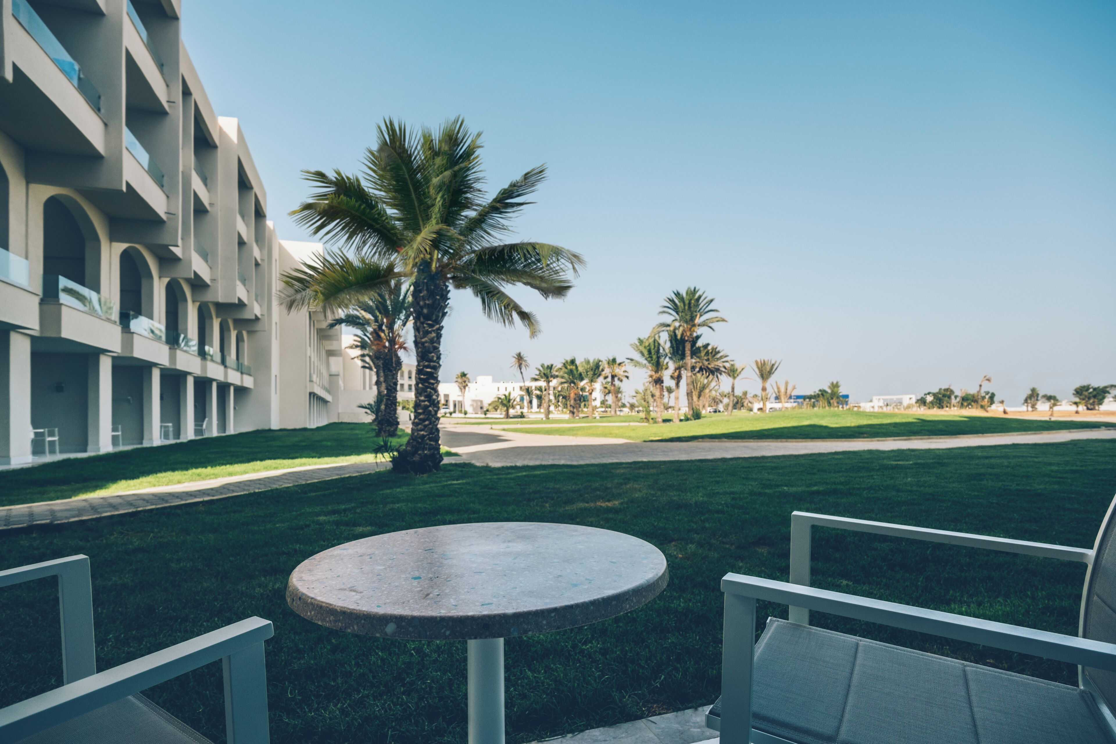 A modern building with a courtyard, palm trees, and a round table with chairs.