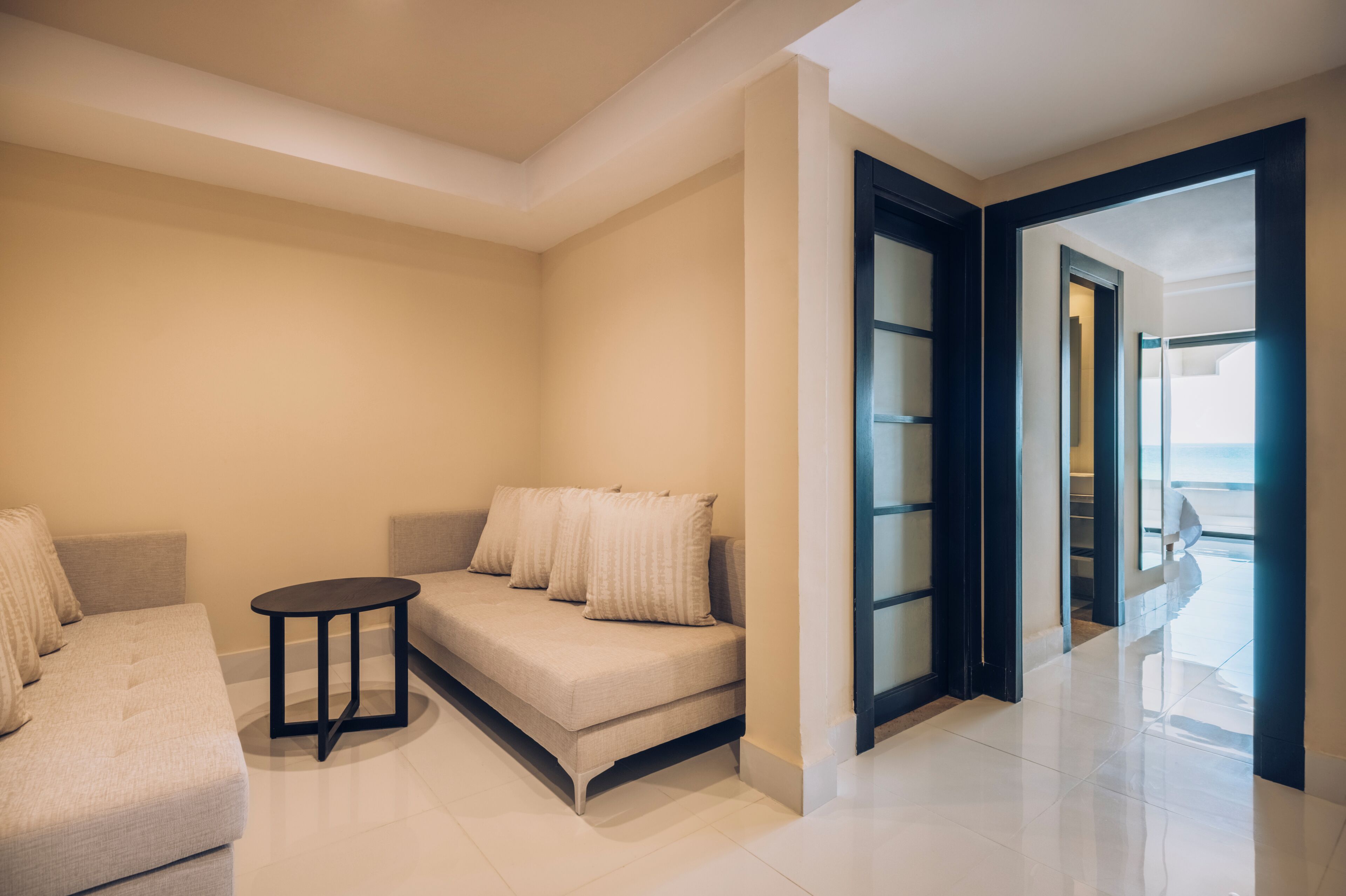 A modern living room with a sofa and a small table, a glass door leading to a balcony, and a view of the sea.