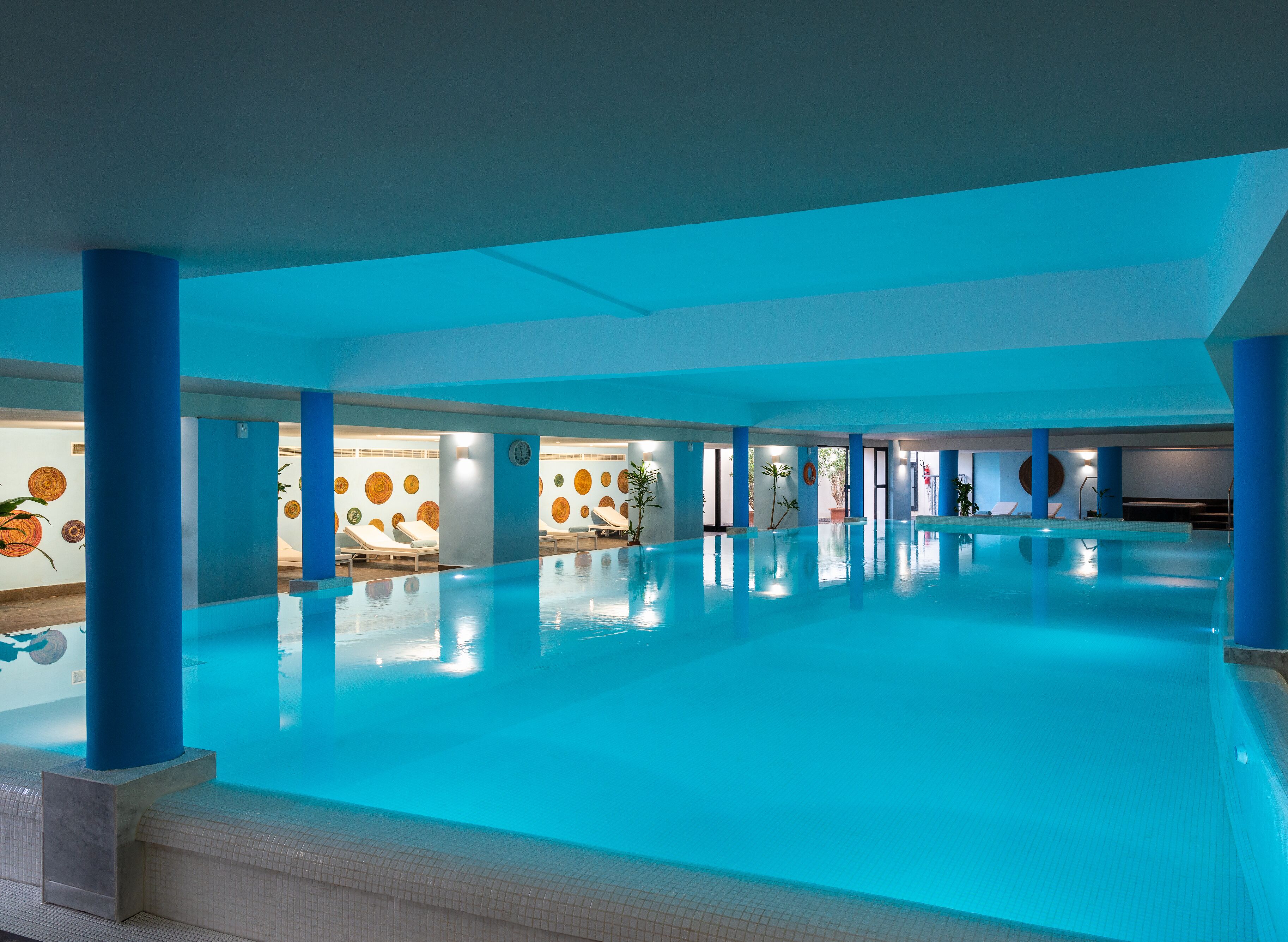 Una amplia piscina interior con iluminación azulada, varias zonas de descanso y decoración artística en las paredes.