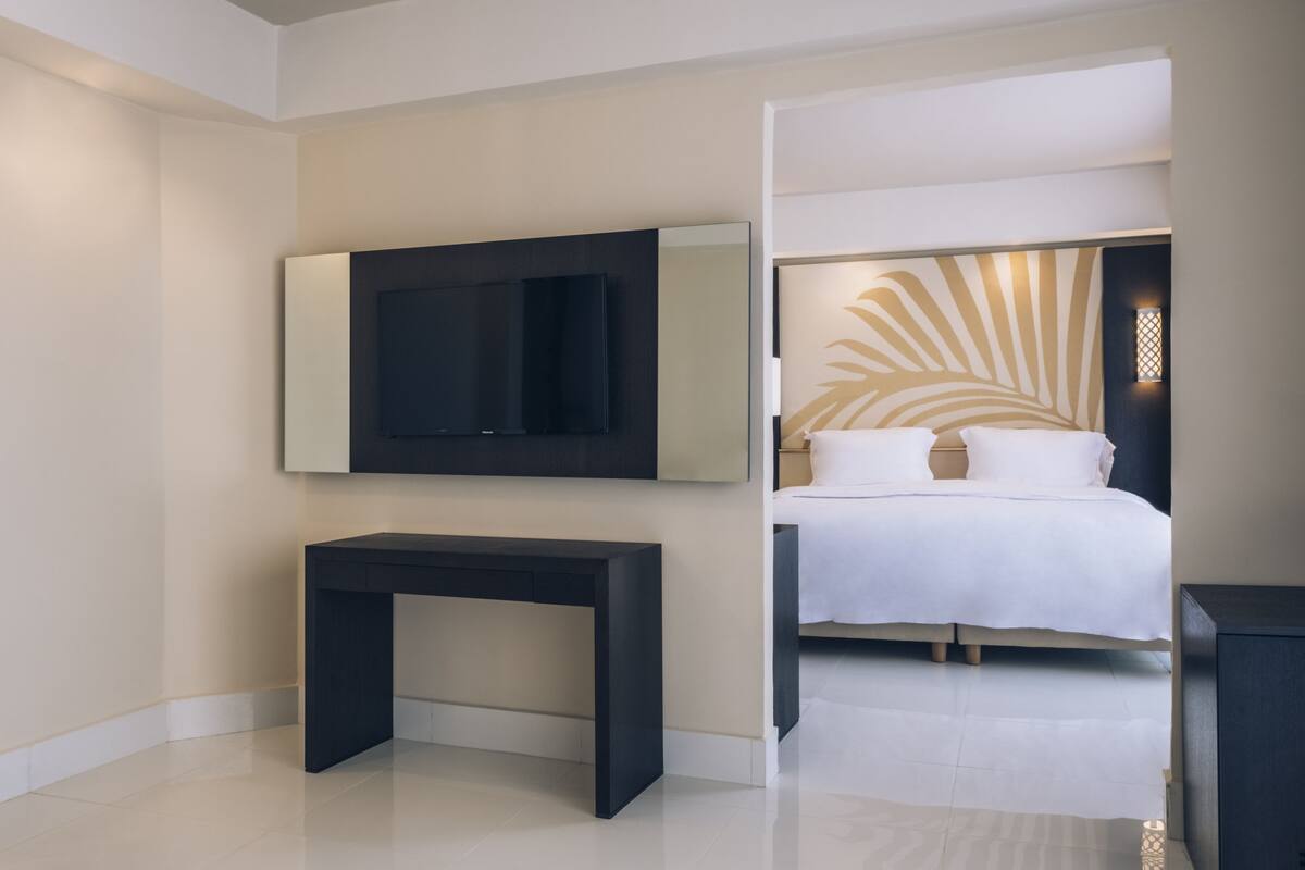 A modern hotel room with a large flat-screen TV mounted on the wall, a dark console table, a neatly made bed with white linens, and a decorative wall panel with palm leaf patterns.