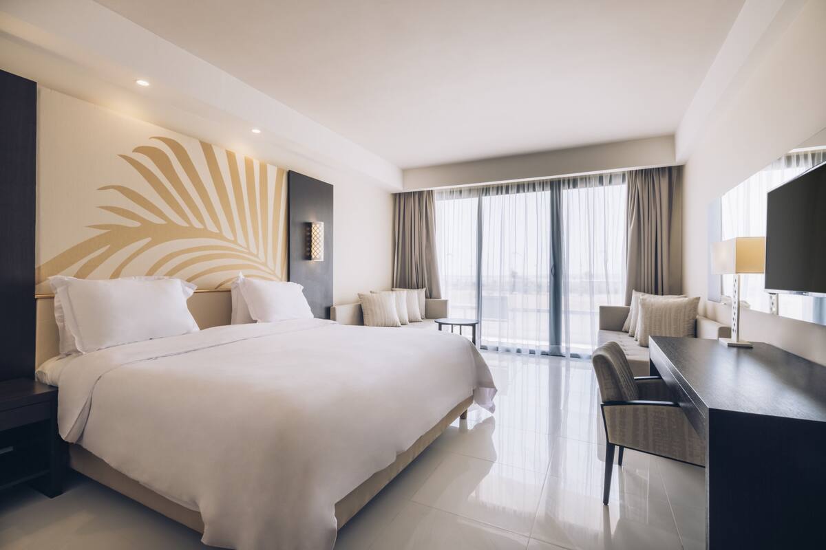 A modern hotel room with a large bed, a desk, and a view of the outdoors through large windows.