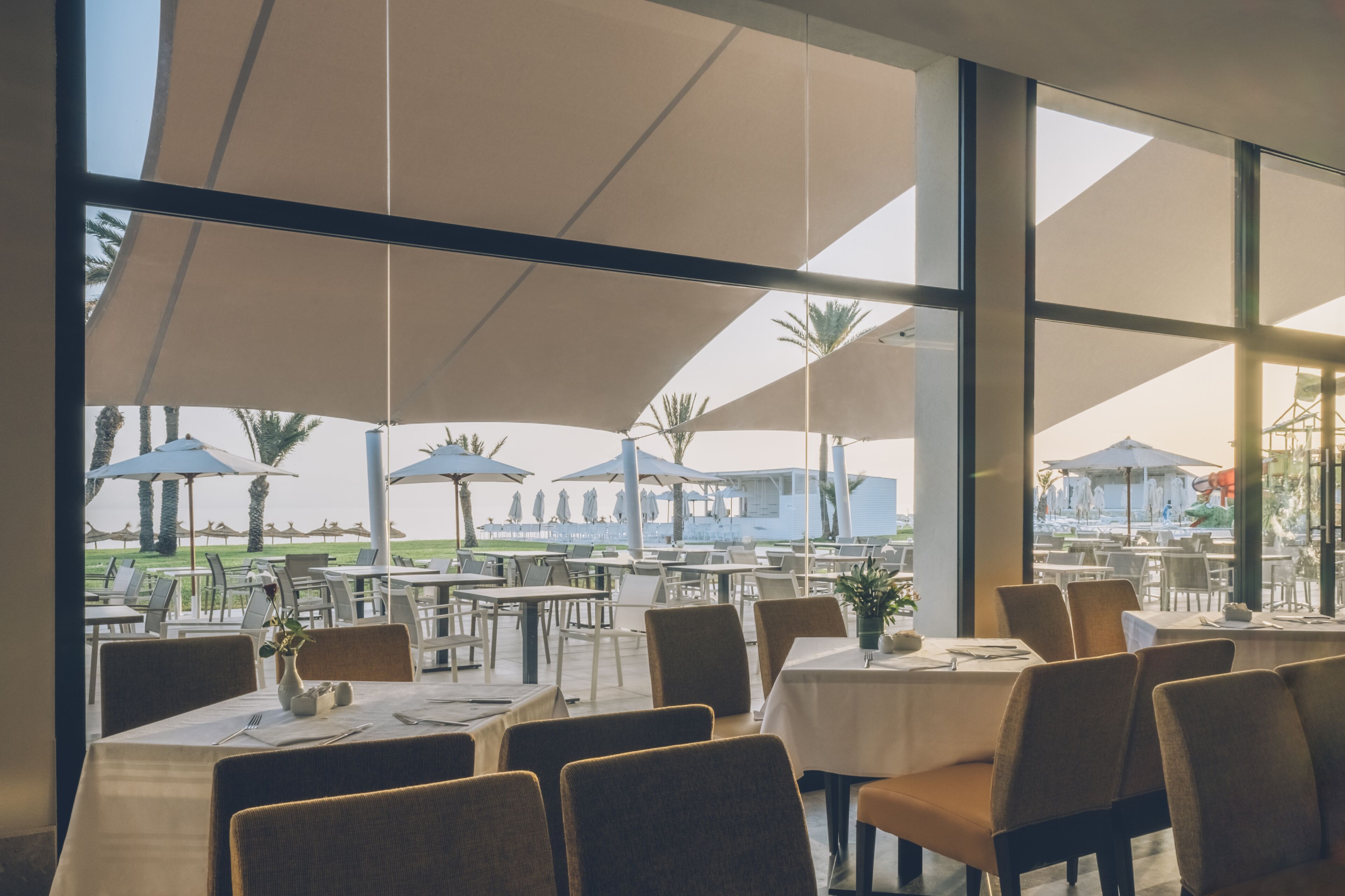 An outdoor dining area with tables and chairs, large umbrellas, and a view of a pool and palm trees.