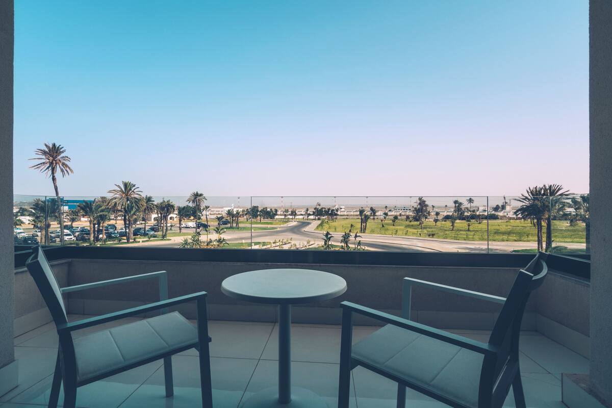 A balcony with two chairs and a small table, overlooking a road with palm trees and a clear sky.