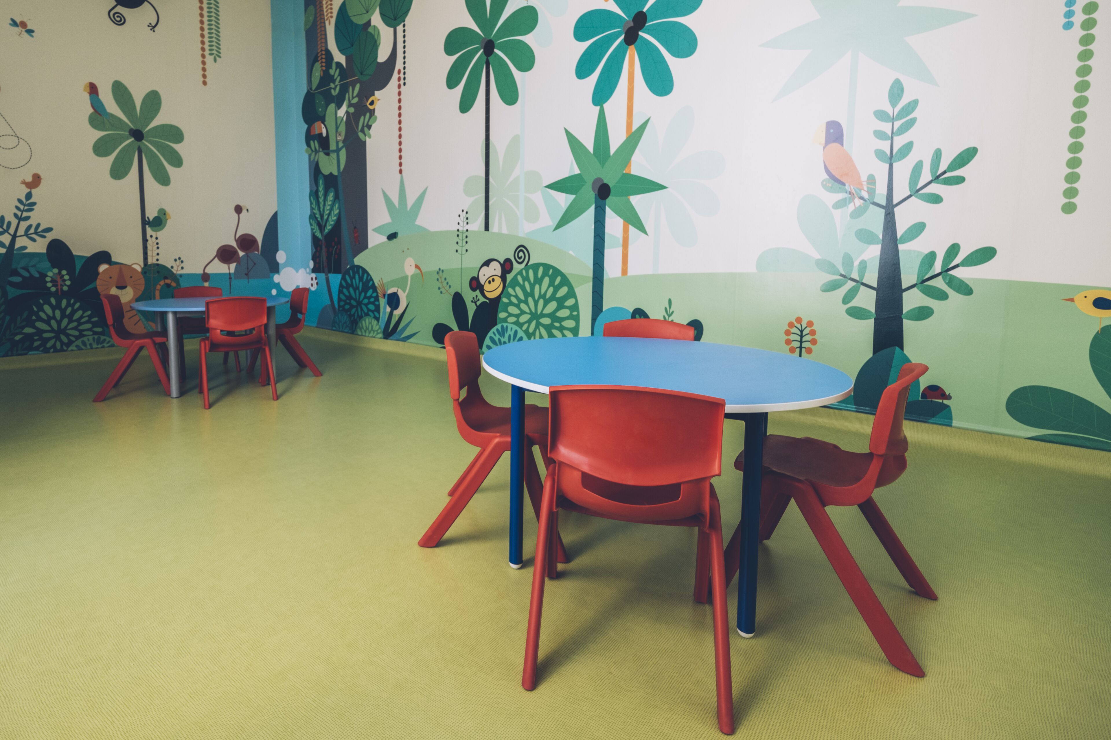 Ein Zimmer mit rundem Tisch und roten Stühlen, einer Wandmalerei mit tropischen Pflanzen und Tieren sowie einem grünen Teppich.