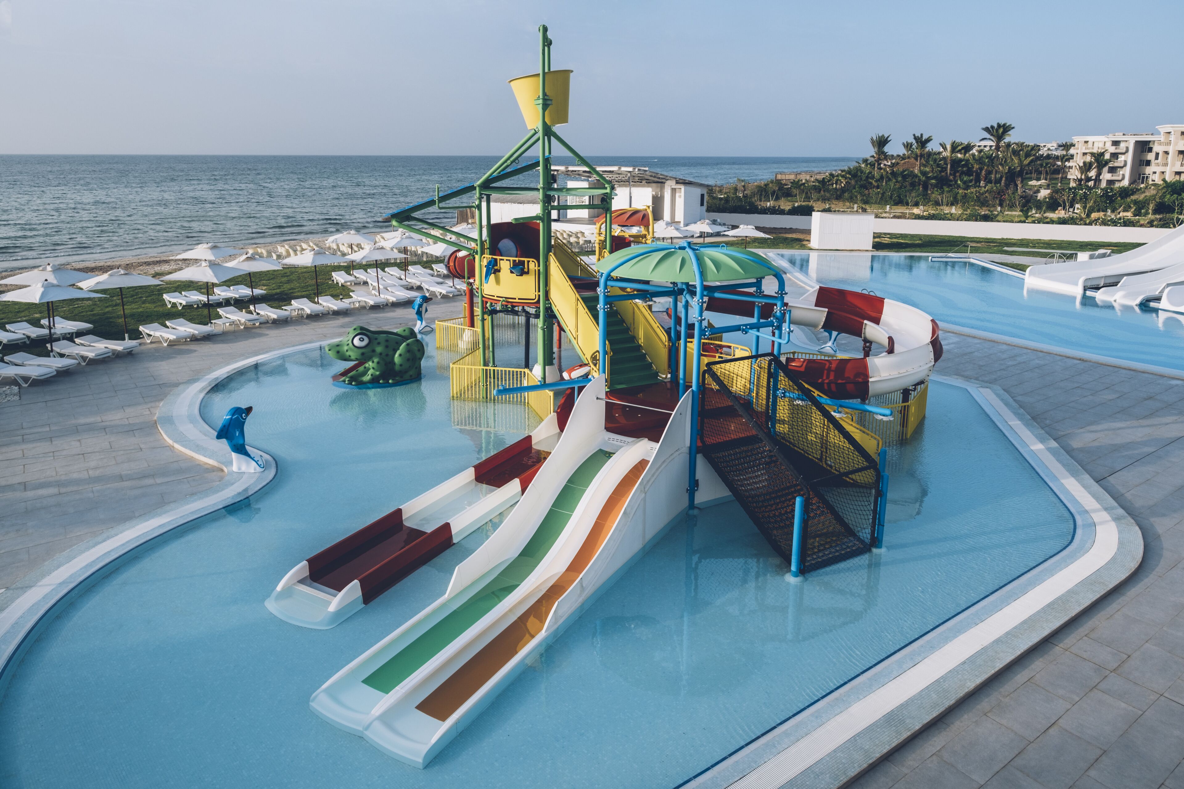 A water park with slides and a play structure near a pool, overlooking a beach and ocean.