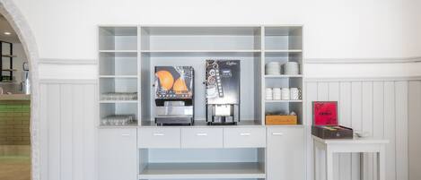Una estación de café blanca, montada en la pared, con dos máquinas de espresso, un expositor de vidrio y una pequeña mesa con una taza de café y una caja de bombones Delica.
