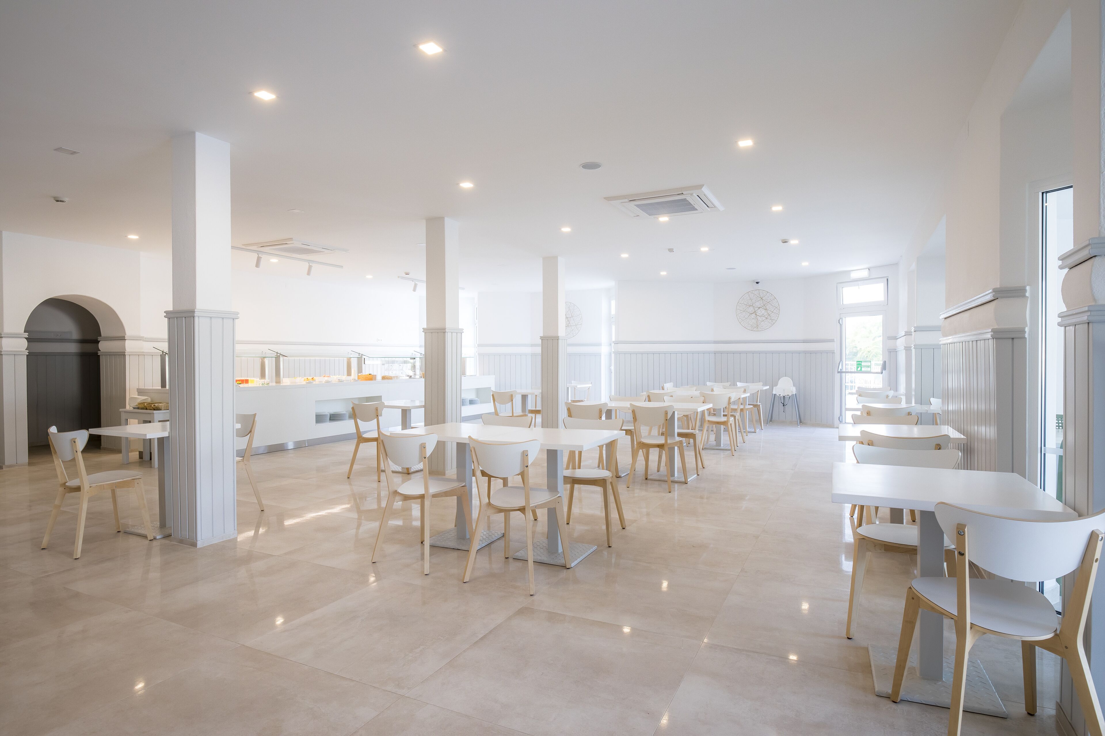 A spacious dining area with white tables and chairs, a central serving counter, and a large window providing natural light.