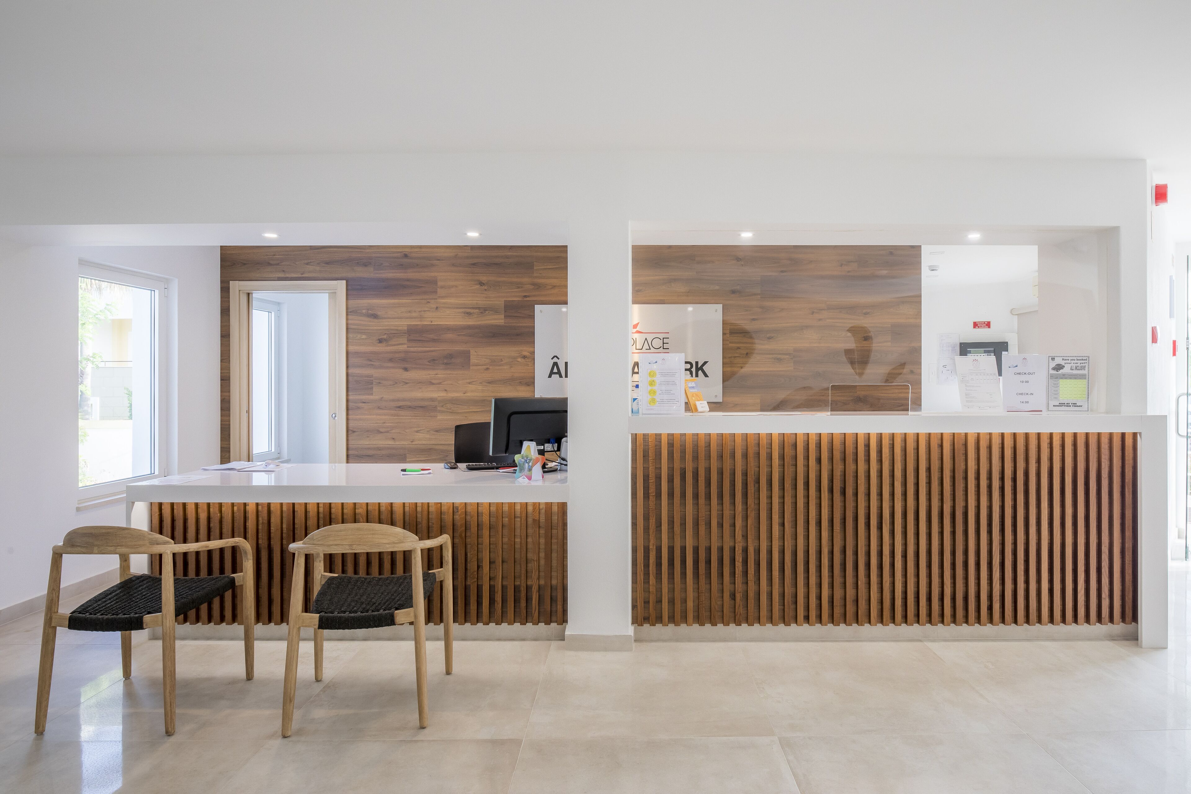 Zona de recepción con un mostrador de madera, dos sillas de madera, una computadora y una ventana con persianas.