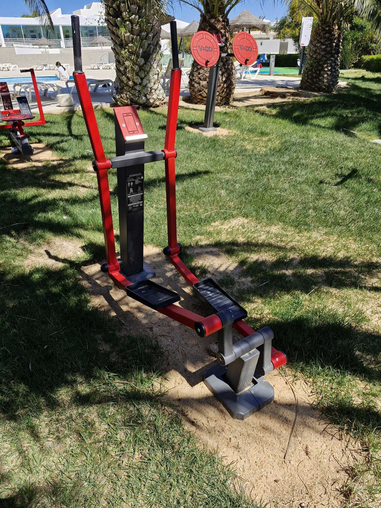 Outdoor exercise equipment with red and black colors, located in a grassy area with palm trees and a pool in the background.