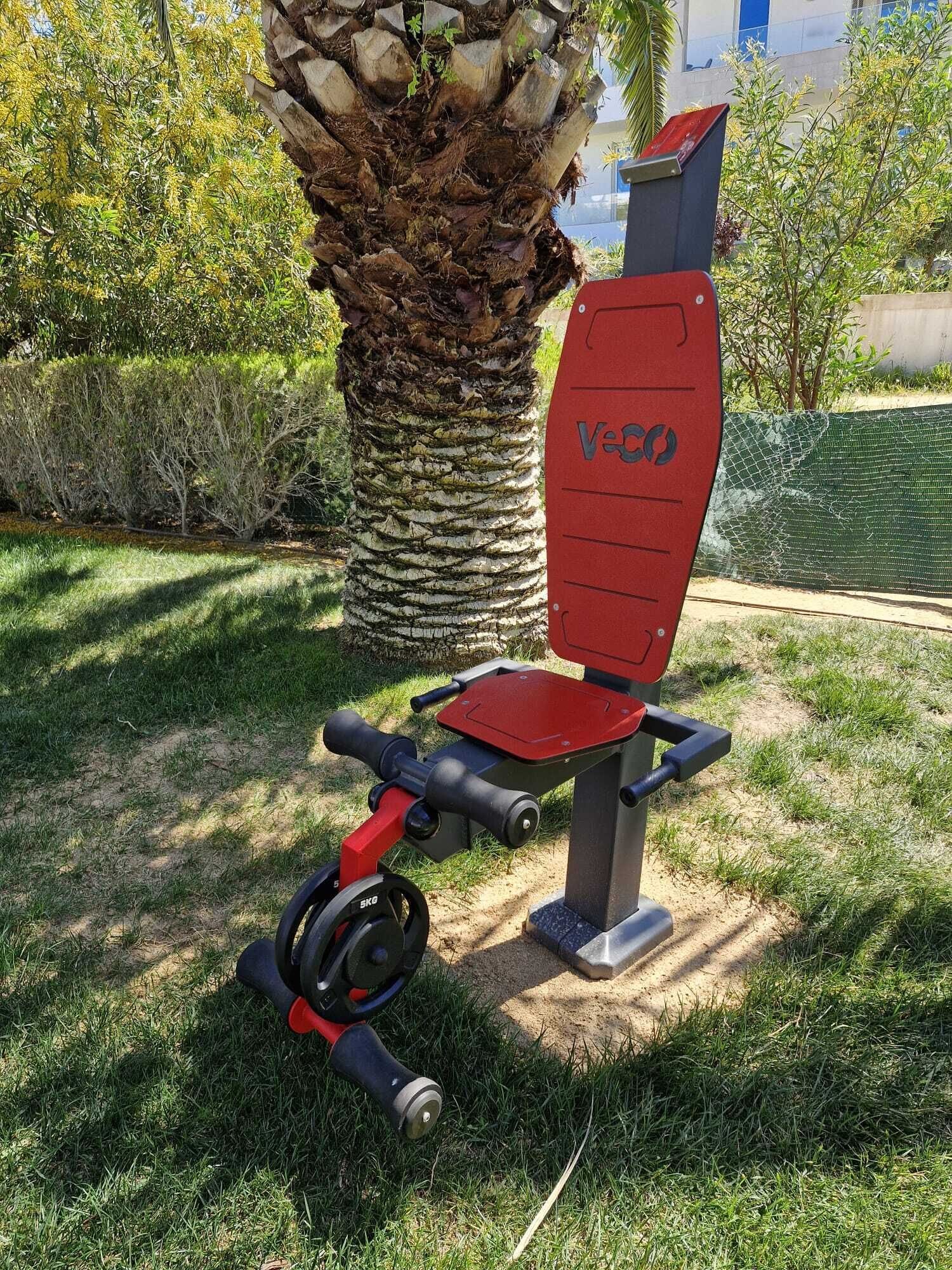 Una bicicleta estática con asiento y manillares rojos, ubicada sobre césped con una palmera y un área con red verde al fondo.
