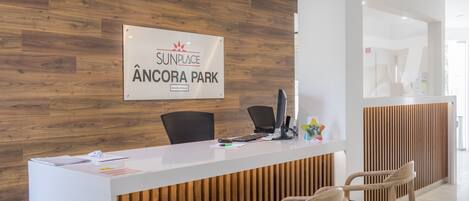 A reception desk with a wooden panel wall, a sign reading "Sunplace Âncora Park", and two chairs in front.
