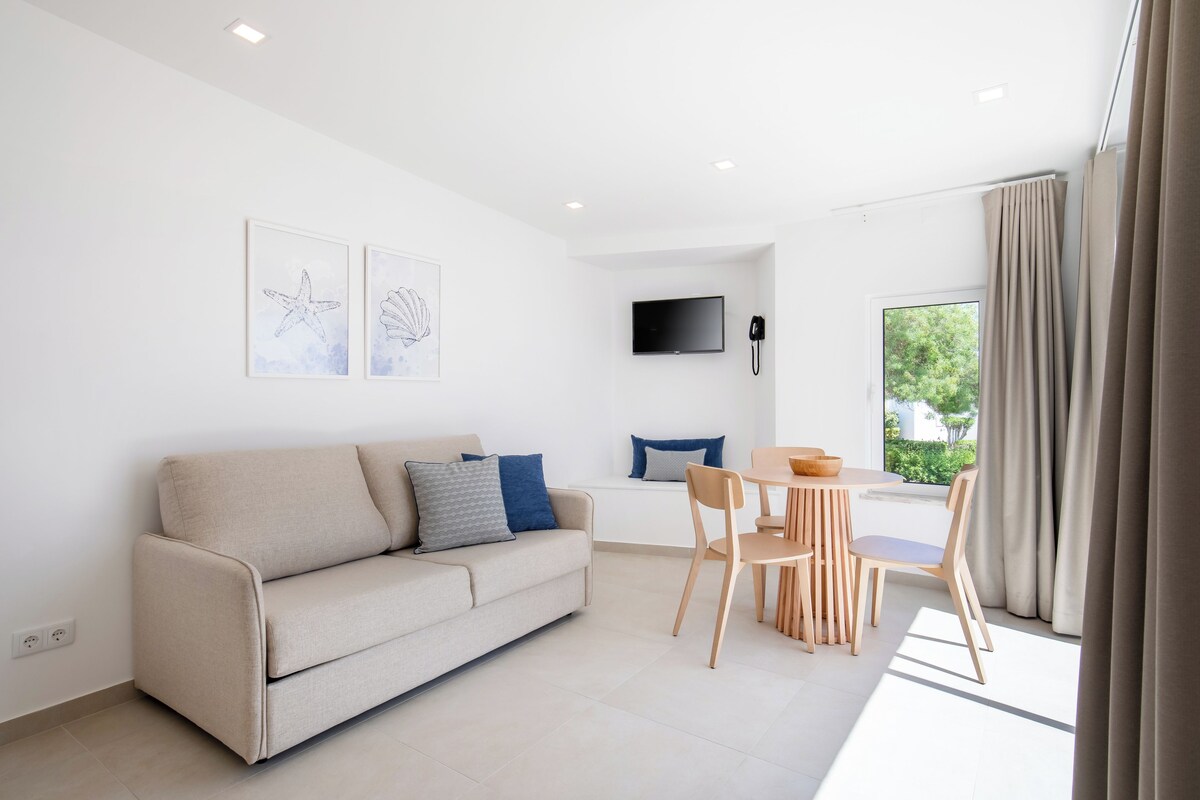 A modern living room with a sofa, a dining table, and chairs. There is a flat-screen TV mounted on the wall and a window with curtains.