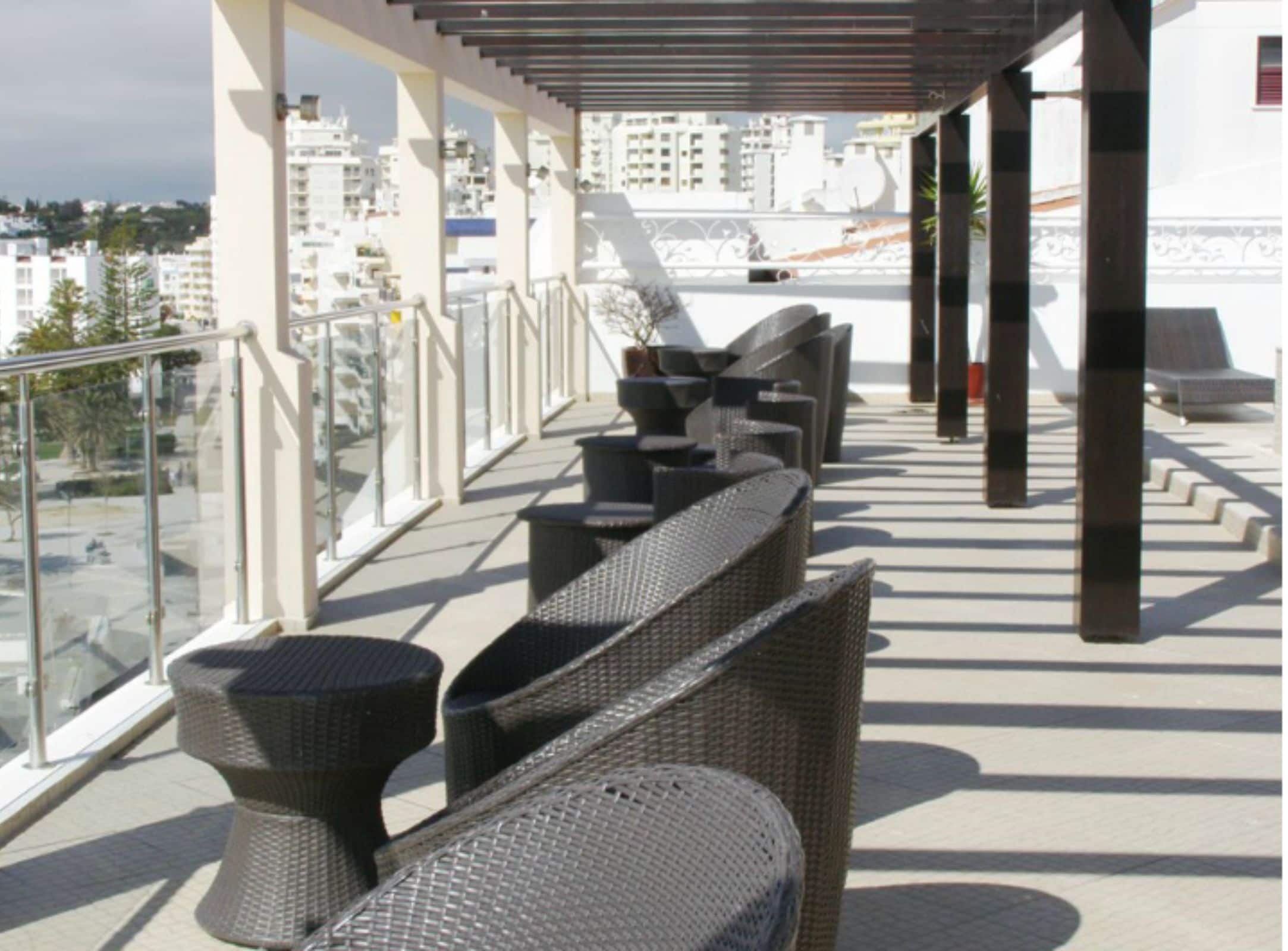 Een terras op het dak met rieten meubilair en een glazen hekwerk, met uitzicht op een stadsgezicht.