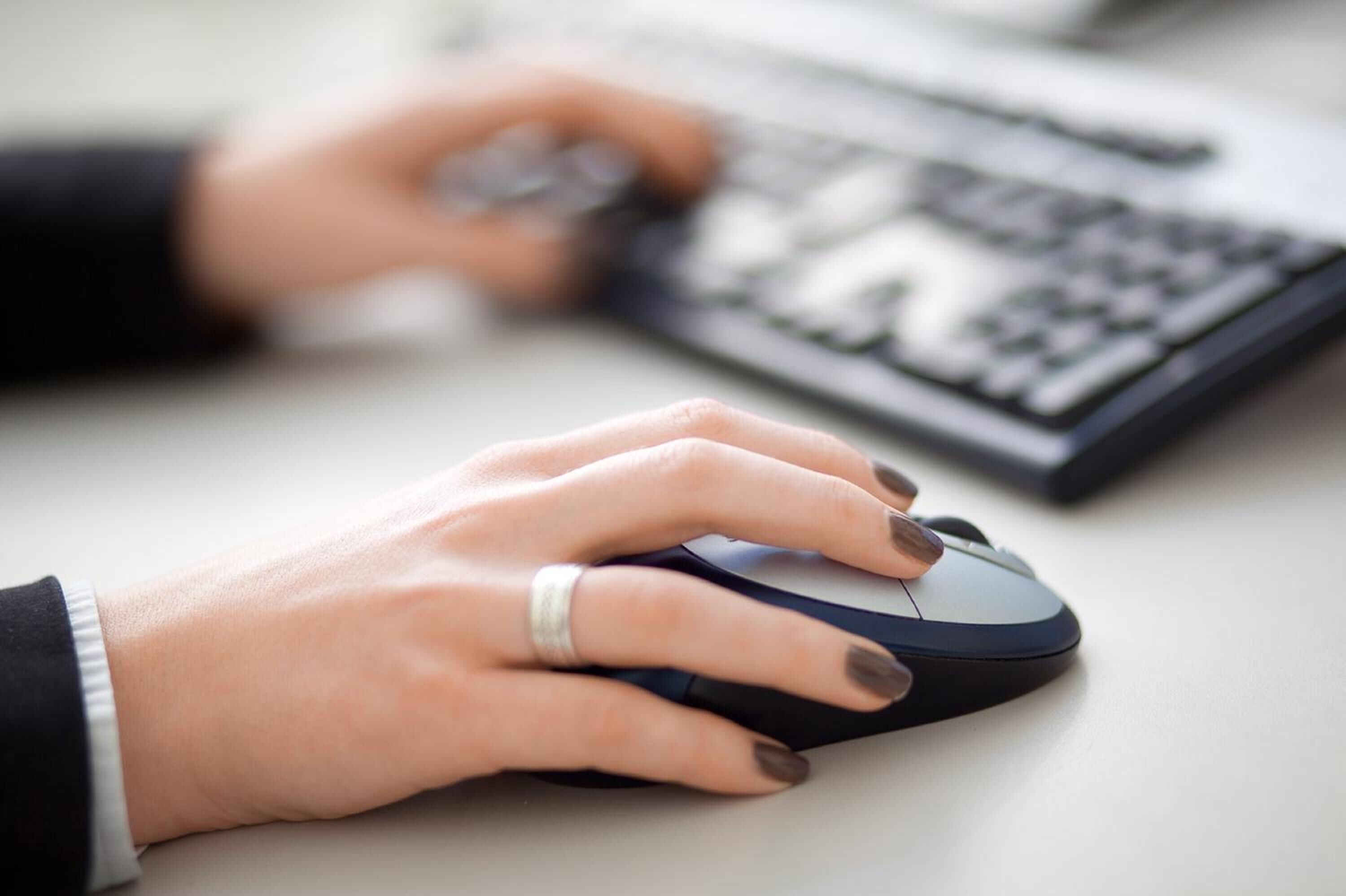 A person's hands using a computer mouse on a desk with a keyboard.
