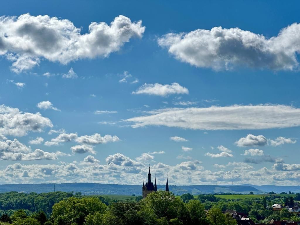 A castle with multiple spires, surrounded by lush green trees and a clear blue sky with scattered clouds.