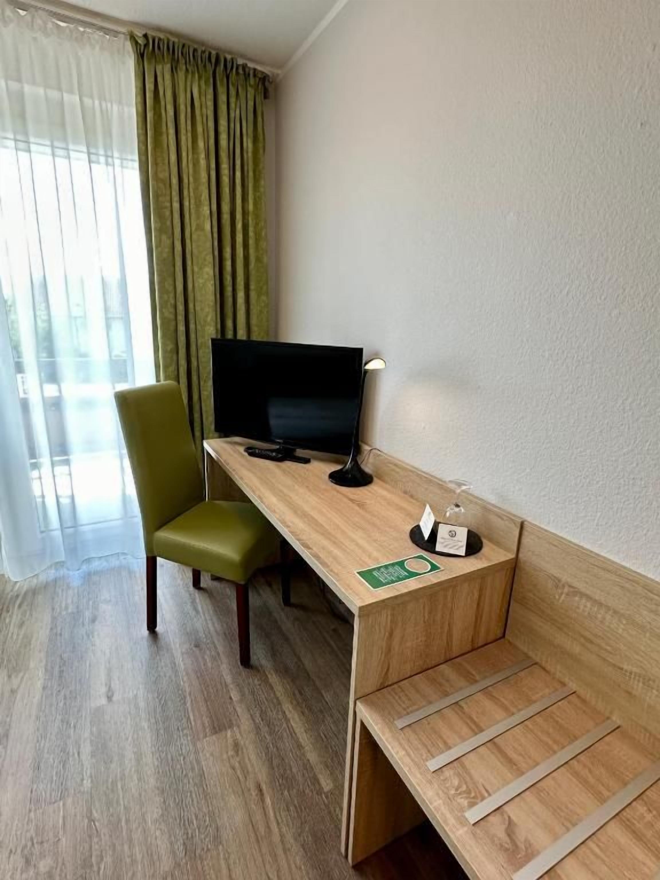 A hotel room with a wooden desk, a green chair, a television, and a window with curtains.