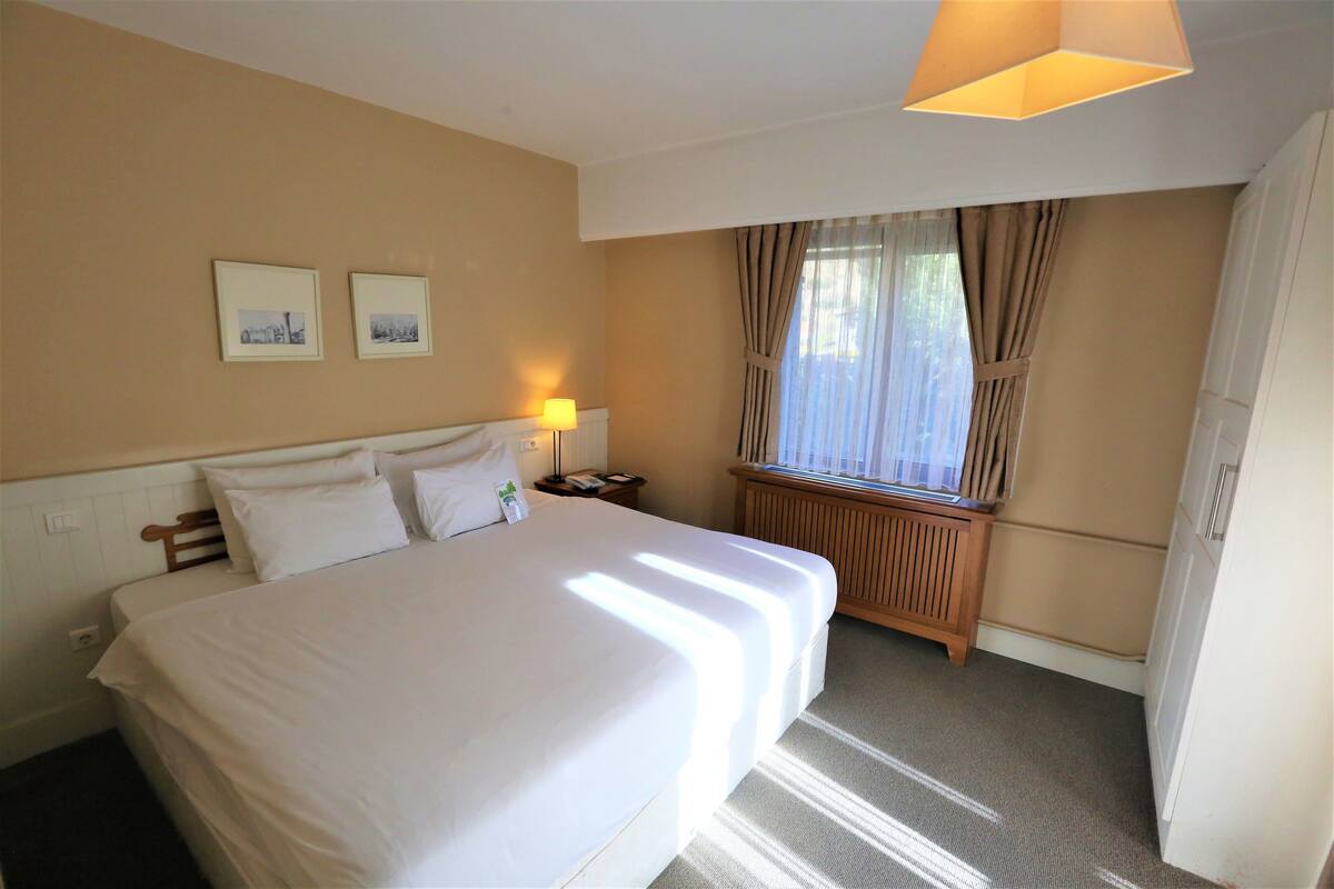 A hotel room with a large bed, a bedside table with a lamp, a window with curtains, and two framed pictures on the wall.