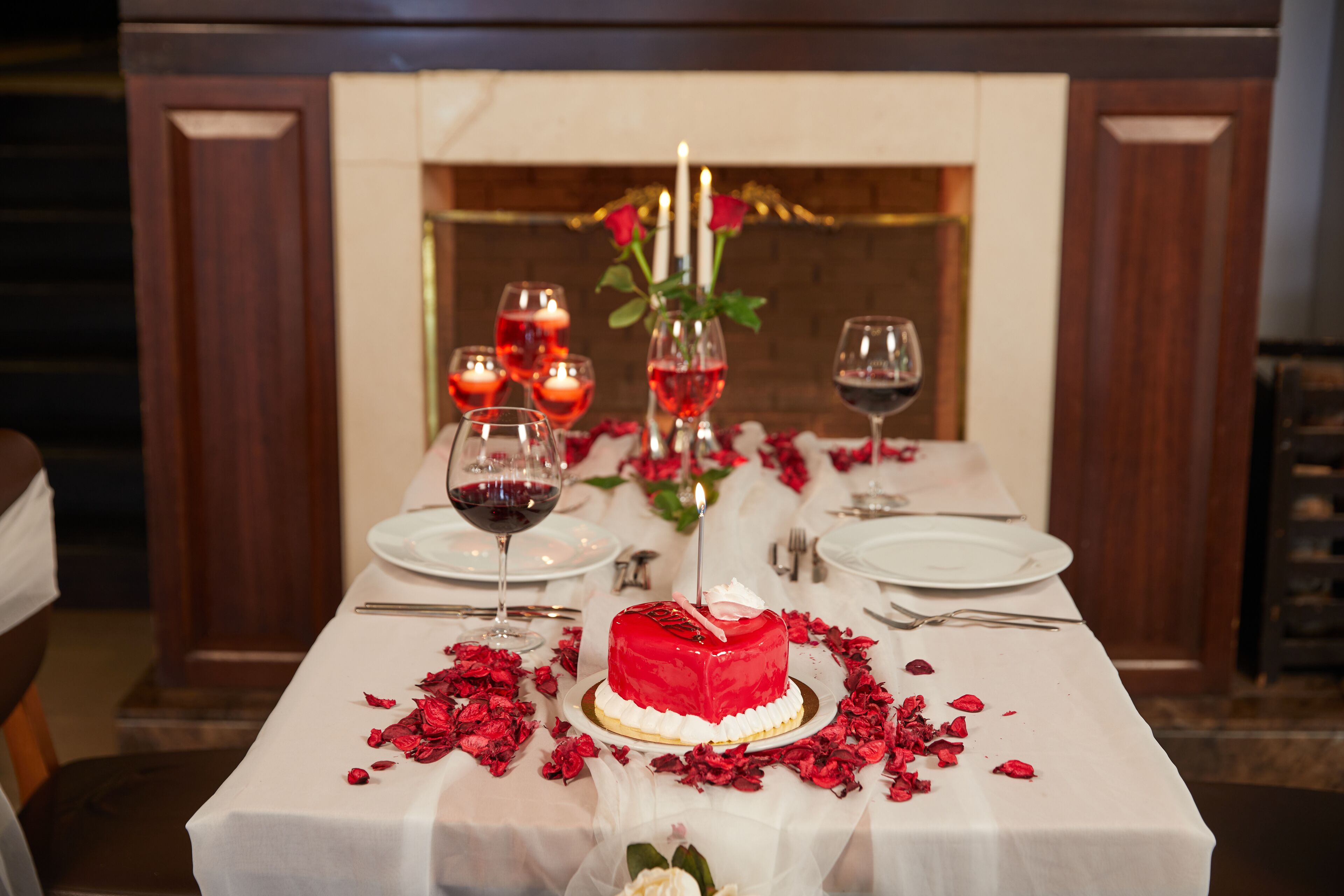 Una mesa dispuesta para una cena romántica, con un pastel en forma de corazón rojo, velas y pétalos de rosa.
