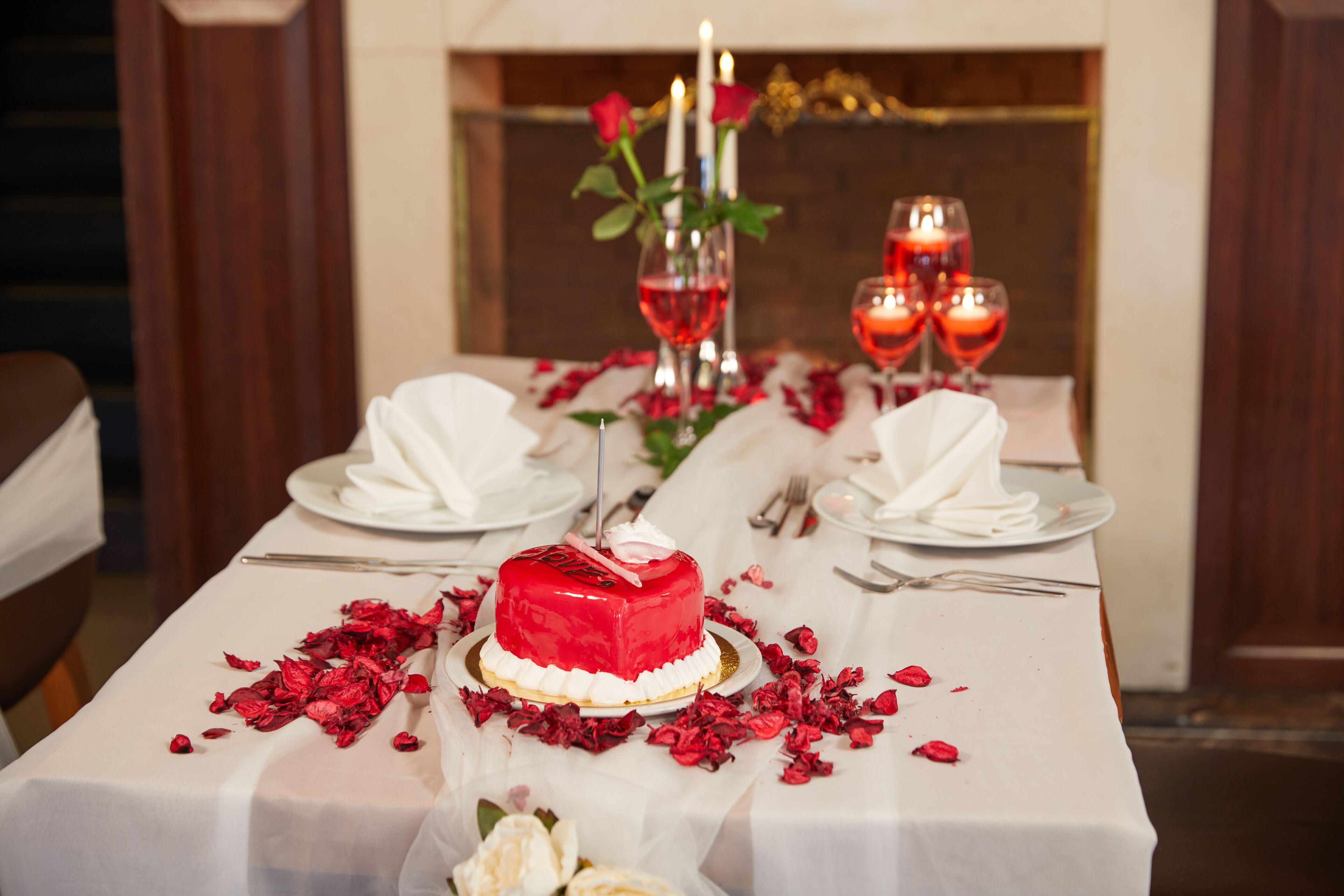 Ein Tisch, festlich für ein romantisches Abendessen gedeckt, mit einer roten Torte, Rosenblättern, Kerzen und Weingläsern.