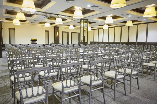 Un salón de conferencias con filas de sillas, un escenario y una pantalla para proyector.