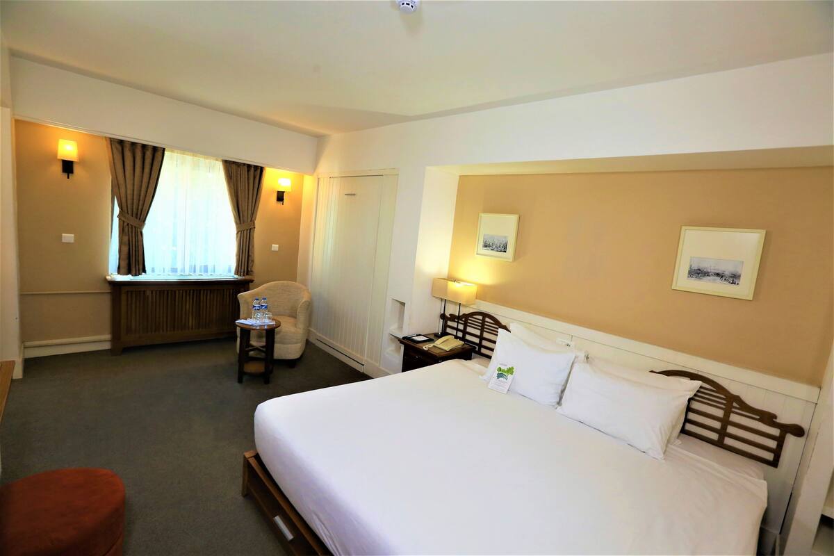 A hotel room with a large bed, a small table with a lamp, a chair, and a window with curtains.