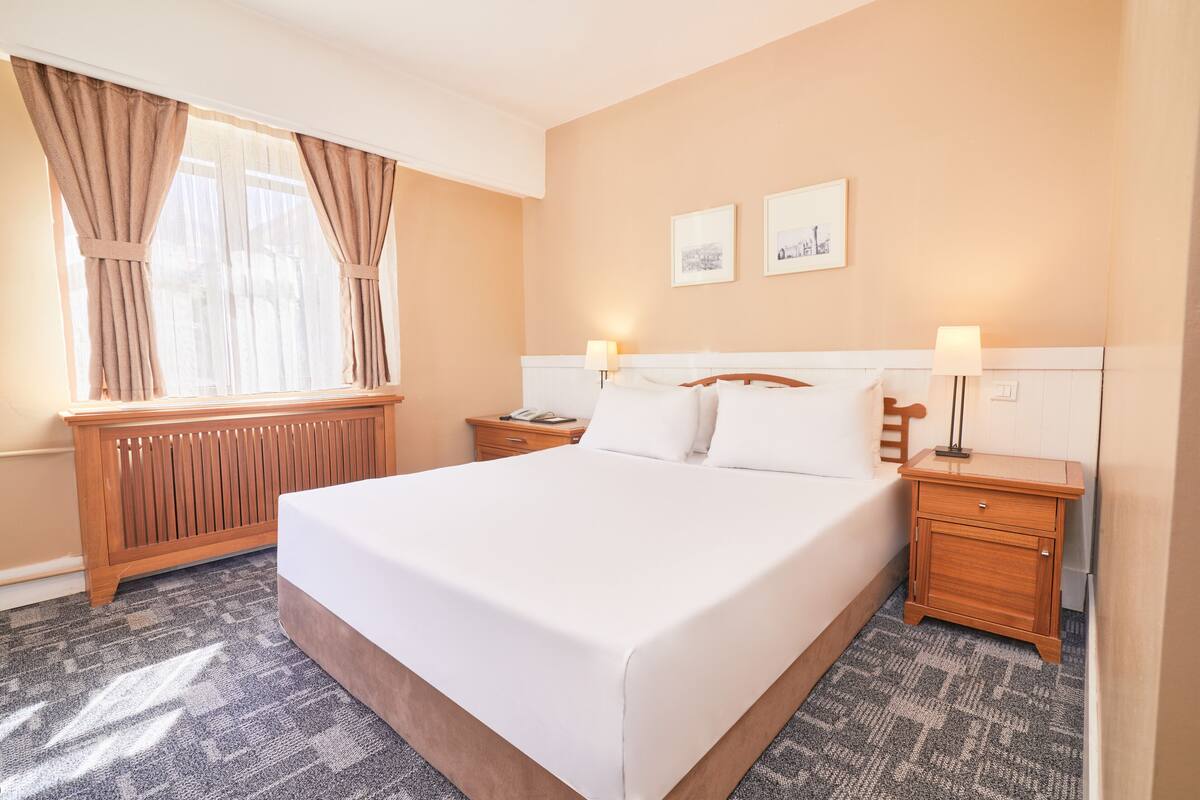 A hotel room with a large bed, two bedside tables, a wooden nightstand, a radiator, and a window with curtains.