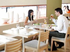 A restaurant with wooden chairs and tables, a window with a view, and a person holding a glass. - Crown Promenade Melbourne (Melbourne)
