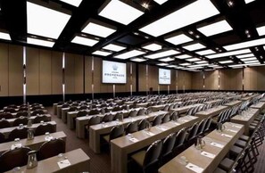 Une salle de conférence avec des rangées de tables et de chaises, un grand écran à l’avant et un projecteur.